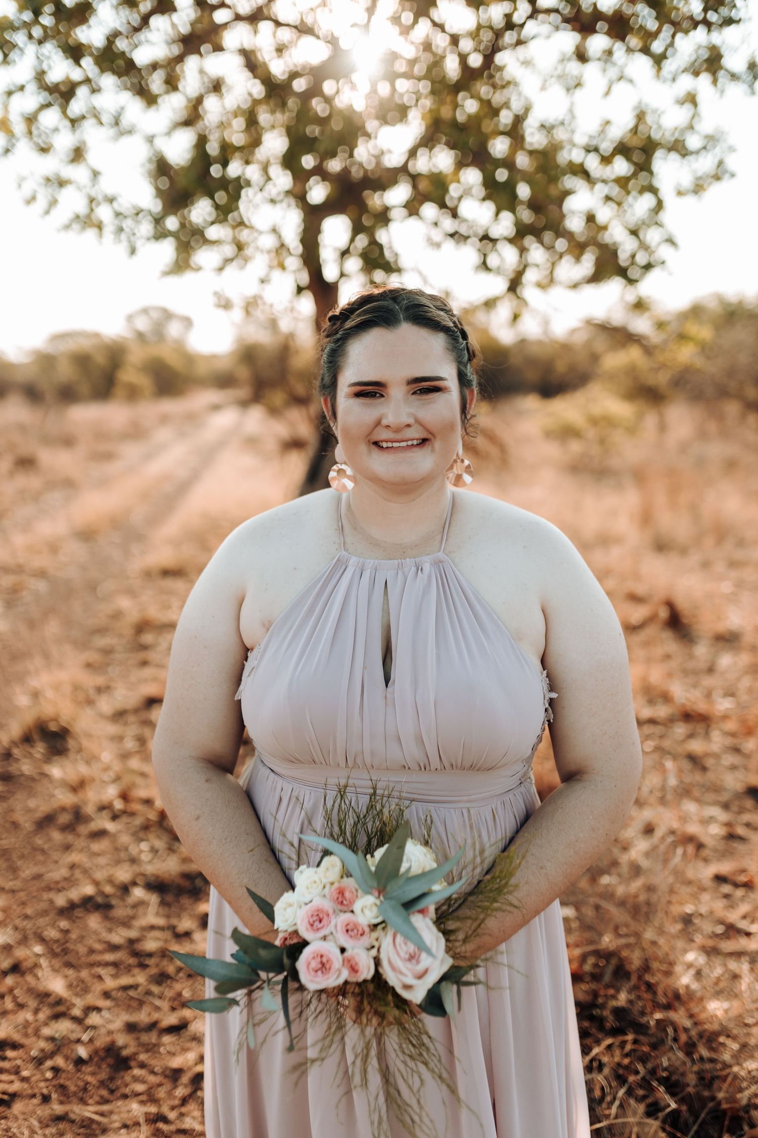 Wedding party member in flowing blush gown holding pastel bouquet in outdoor setting.