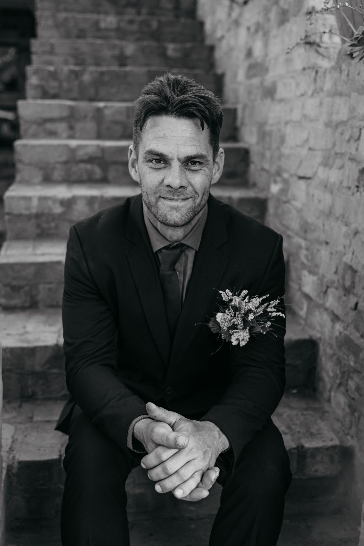 Black and white portrait of a groom sitting on stone steps in a formal suit.