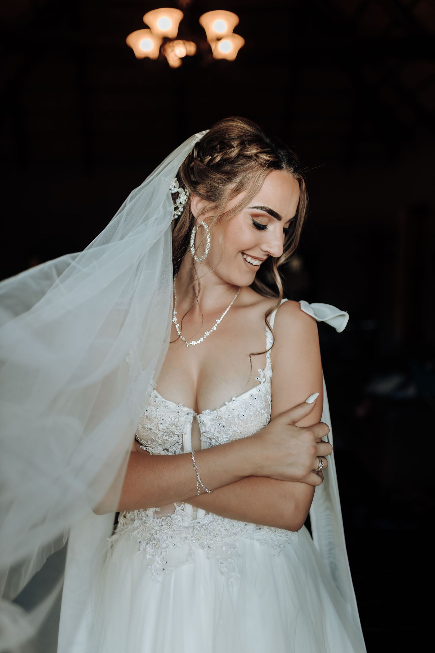 Bride in lace dress with flowing veil poses under warm lighting fixtures.