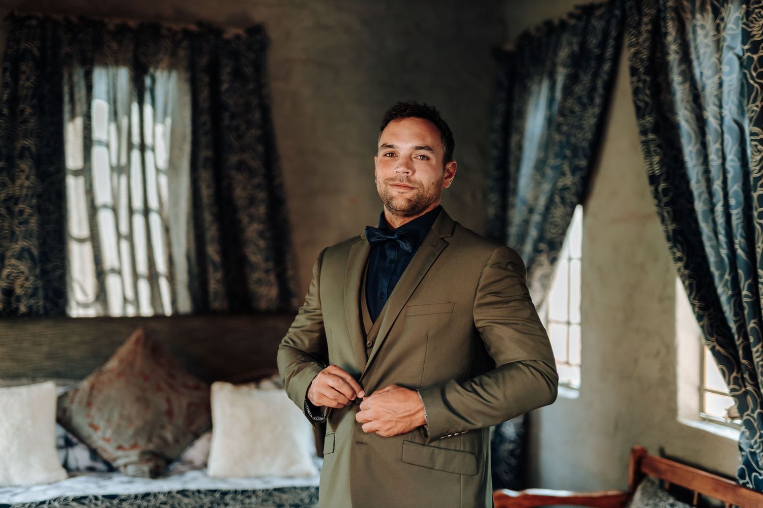 Person wearing tan suit and navy shirt poses in bedroom setting.