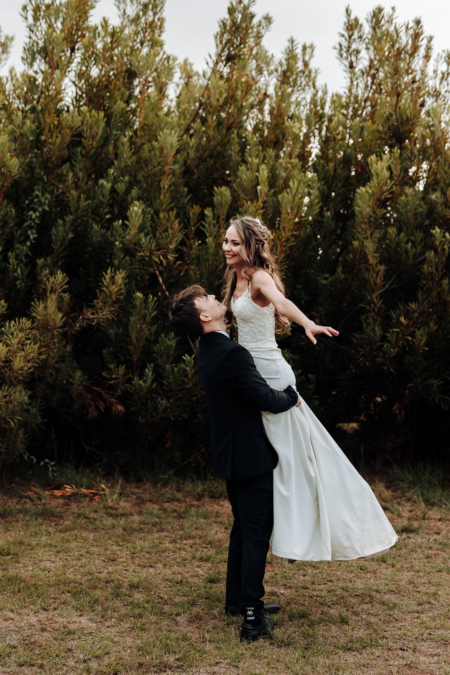 Groom lifts bride in joyful dance pose against backdrop of evergreen trees.