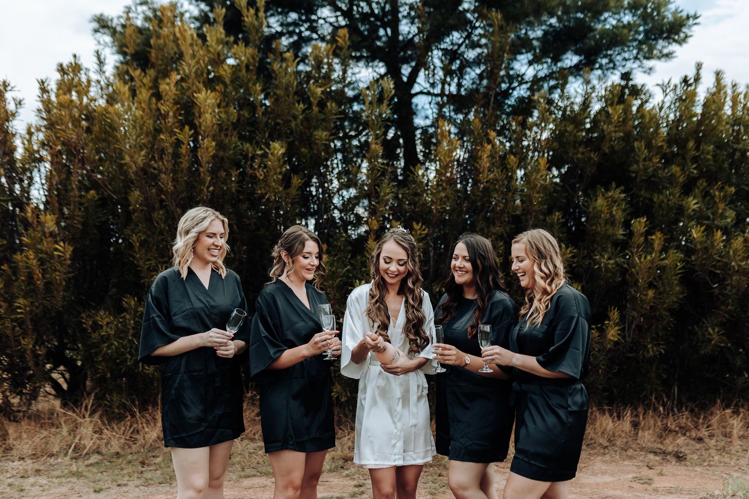 Bridal party in matching robes celebrating outdoors among pine trees.