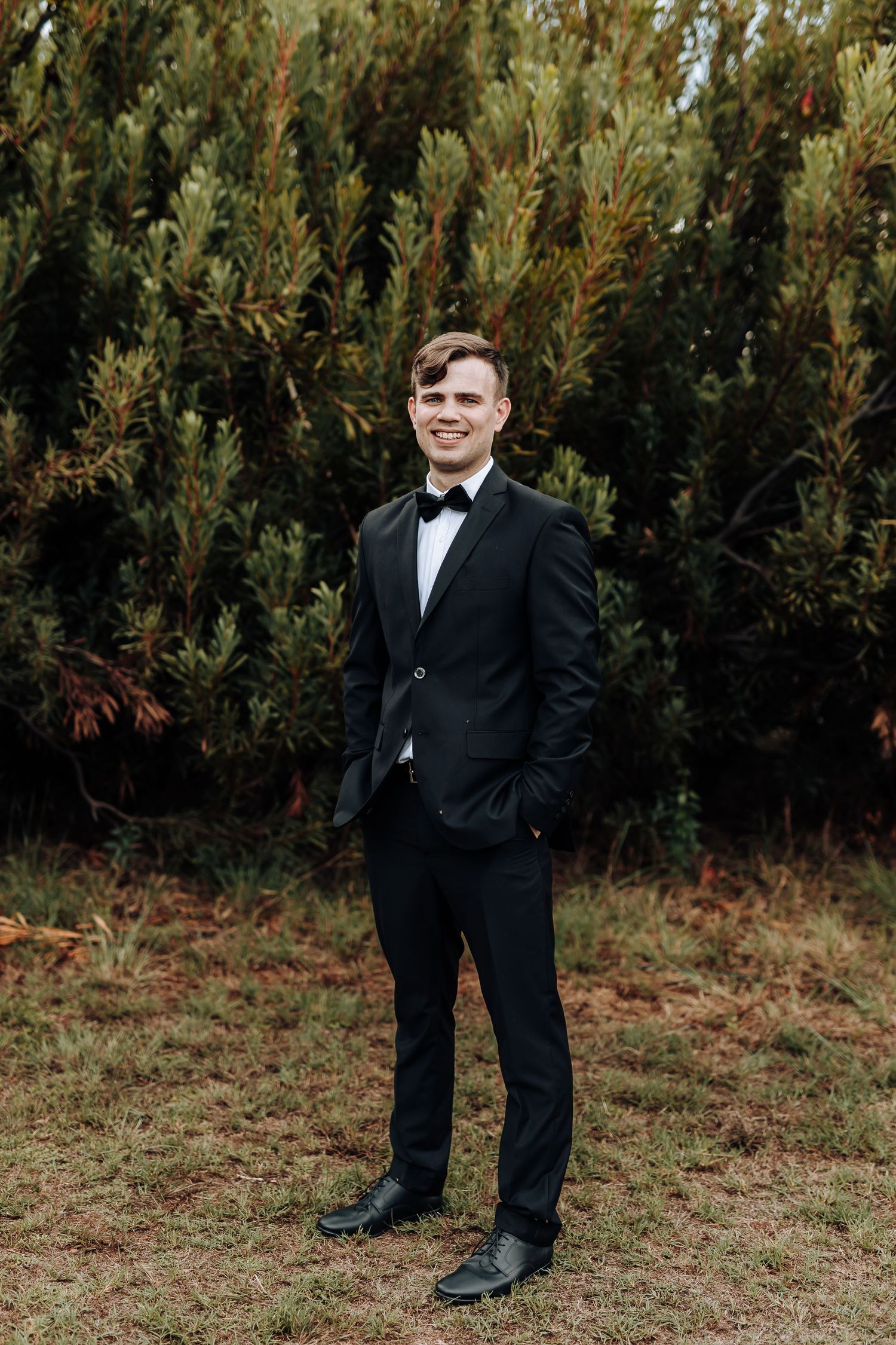 A person in a black tuxedo and bow tie stands in front of evergreen trees.