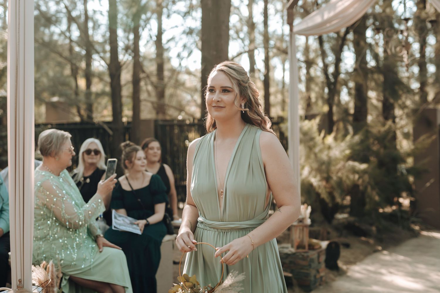 A bridesmaid stands at an outdoor ceremony with guests seated behind her in a wooded setting.