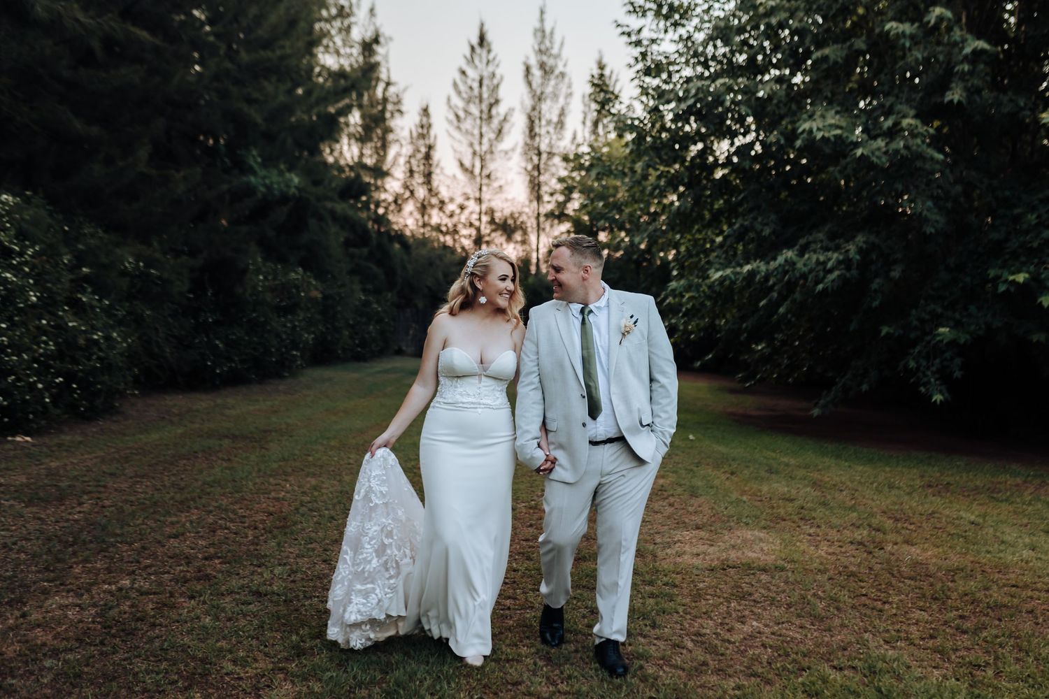 Elegant couple in wedding attire walking through garden path at sunset.