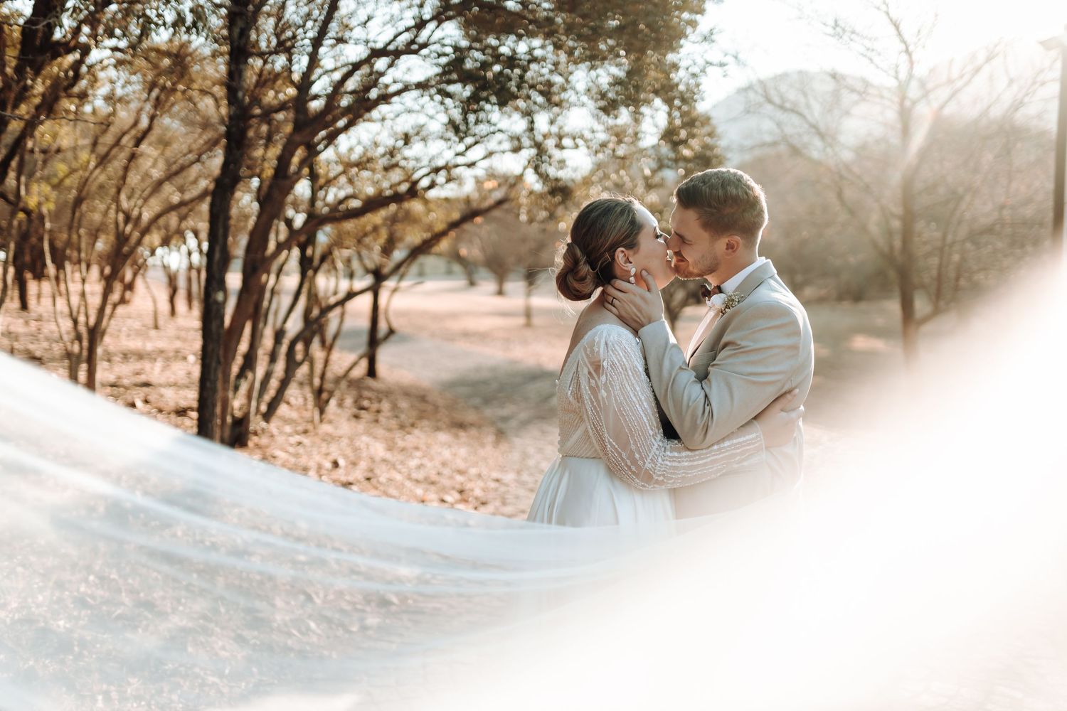 Dreamy outdoor wedding photo with couple embracing among trees and flowing veil.