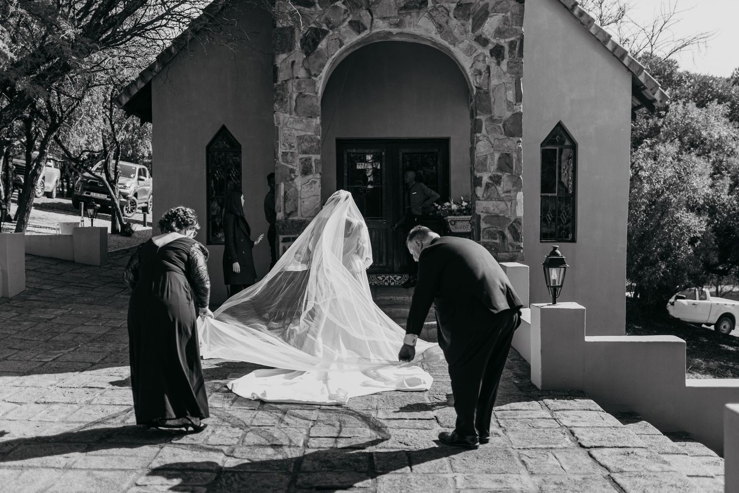 Black and white photo of wedding party adjusting bridal veil outside stone church entrance.