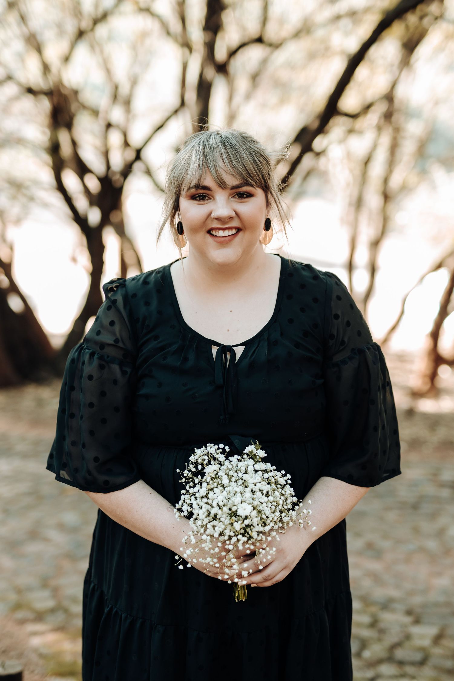 Bridesmaid with bangs wearing black dress smiles while holding white floral bouquet outdoors.
