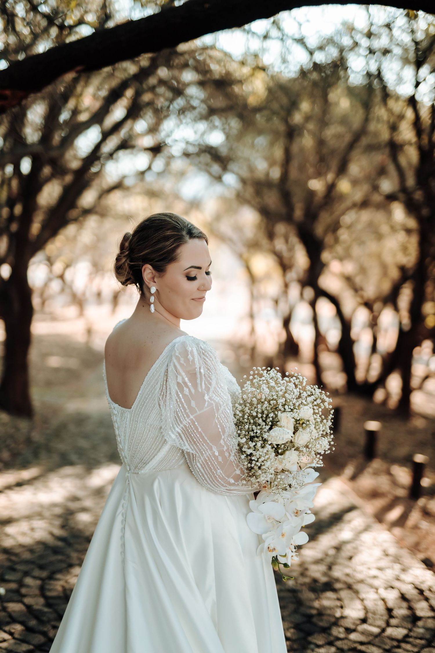 Bride holding white bouquet poses in backless wedding dress on tree-lined cobblestone path.