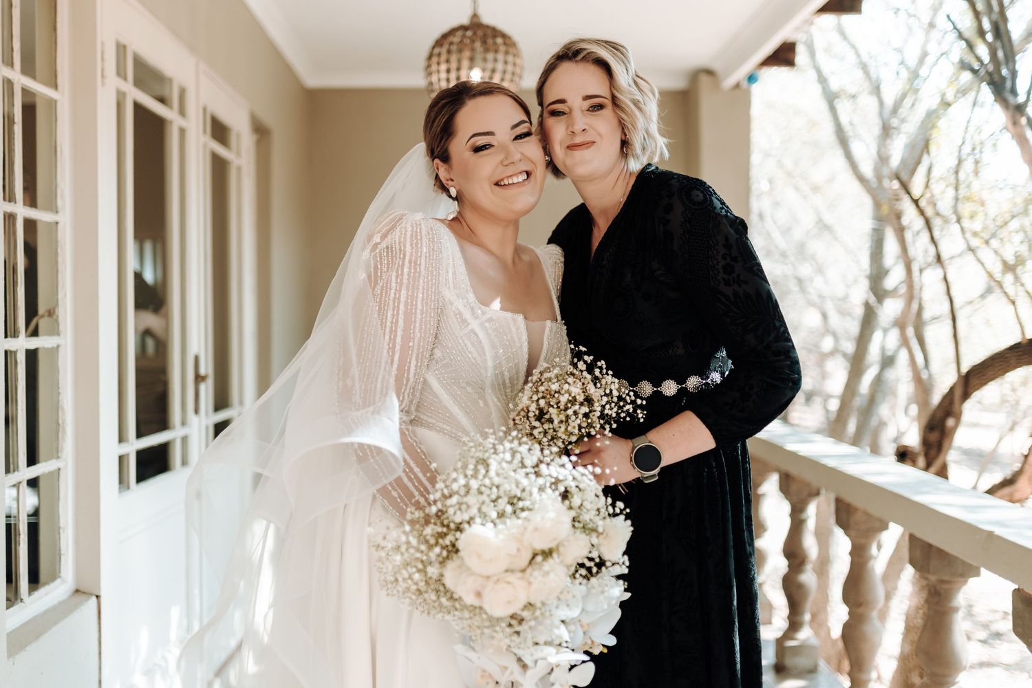 Bride in white gown and bridesmaid in black share intimate moment with bouquets.