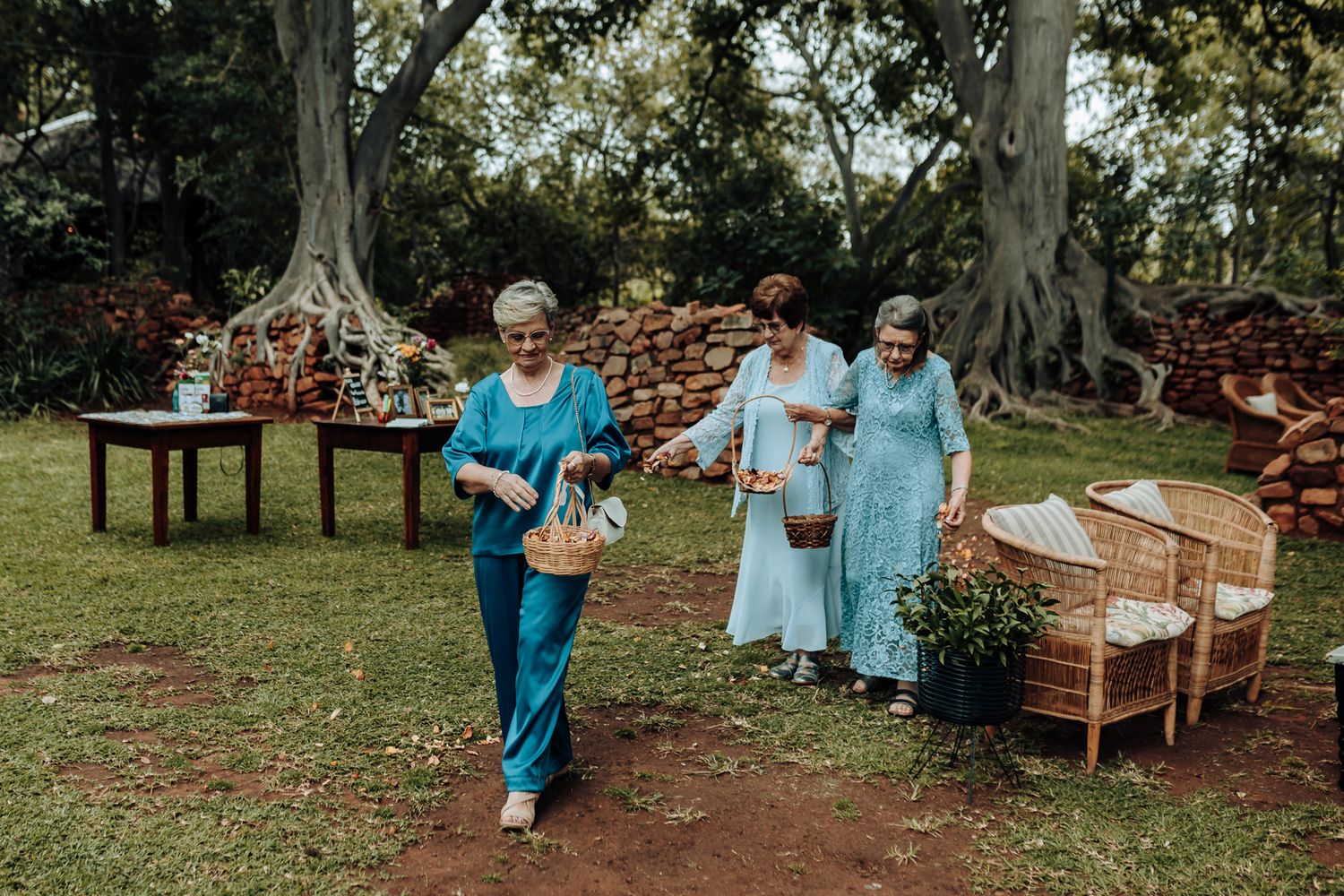 Wedding guests in coordinating blue outfits walk through garden pathway near stone wall.