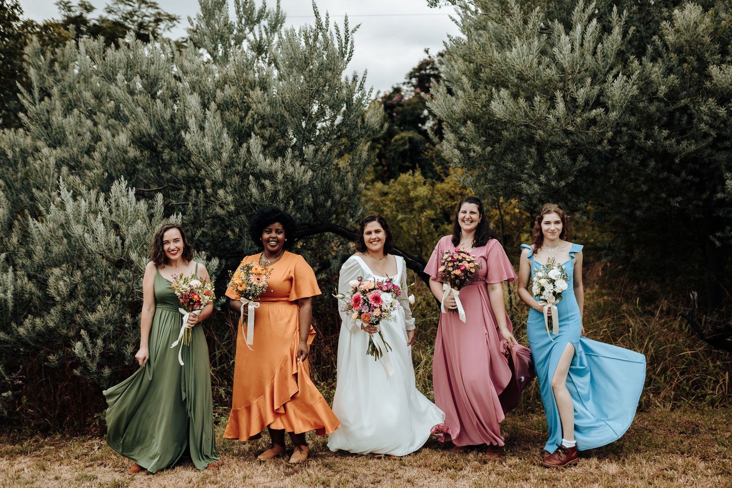 Wedding party in flowing dresses of different colors pose playfully outdoors.