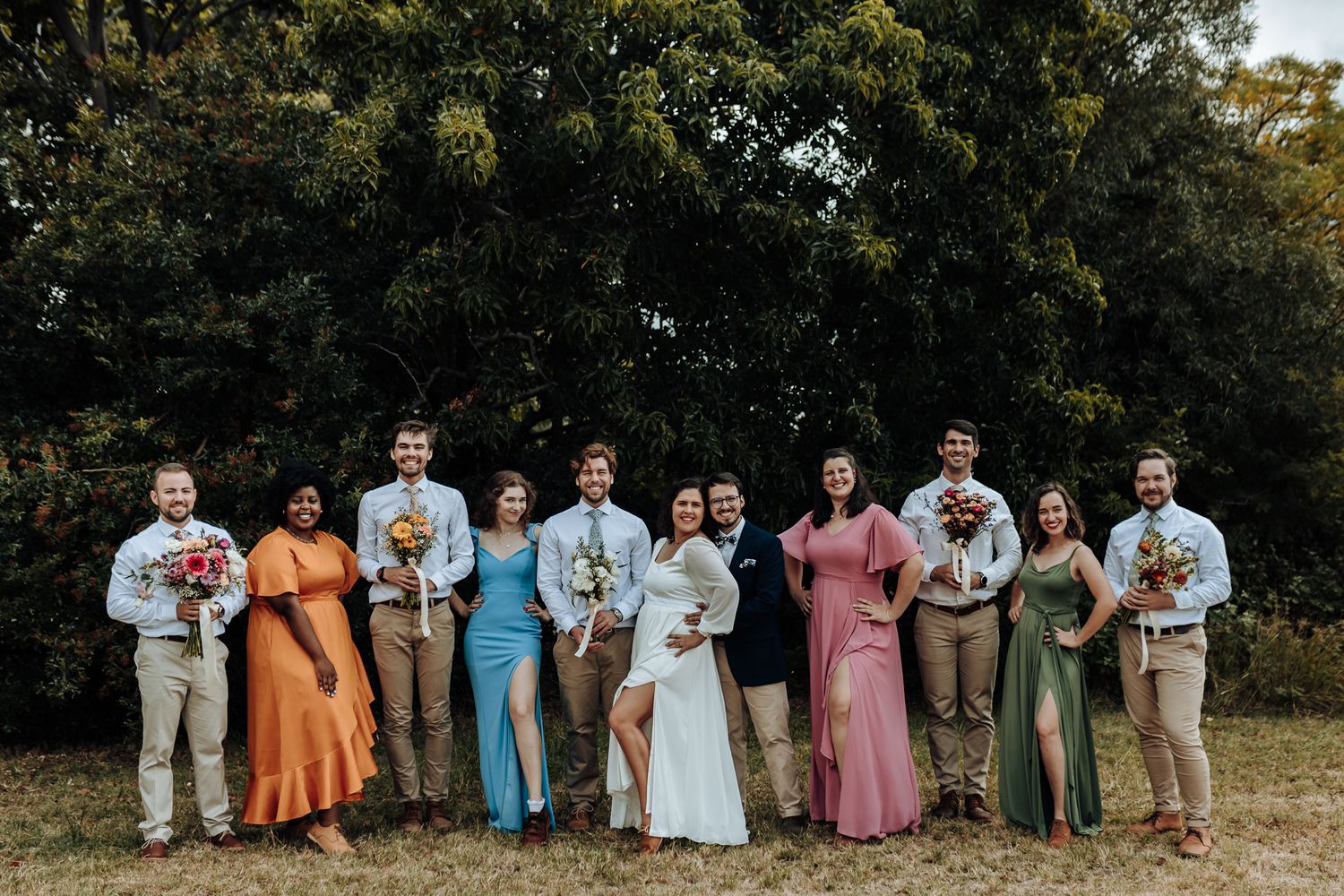 Wedding party in colorful dresses and suits poses together in front of trees.
