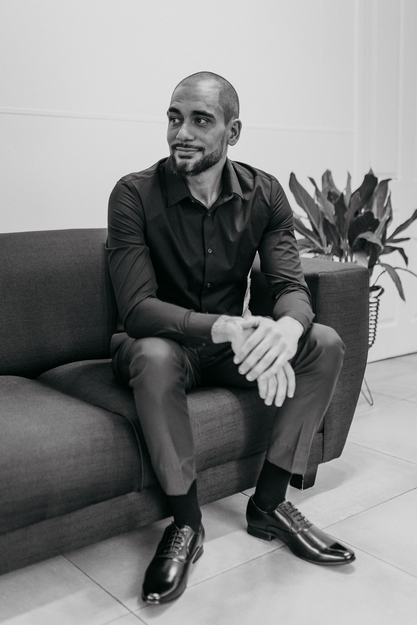 Person seated on grey couch in casual attire with indoor plant in background.