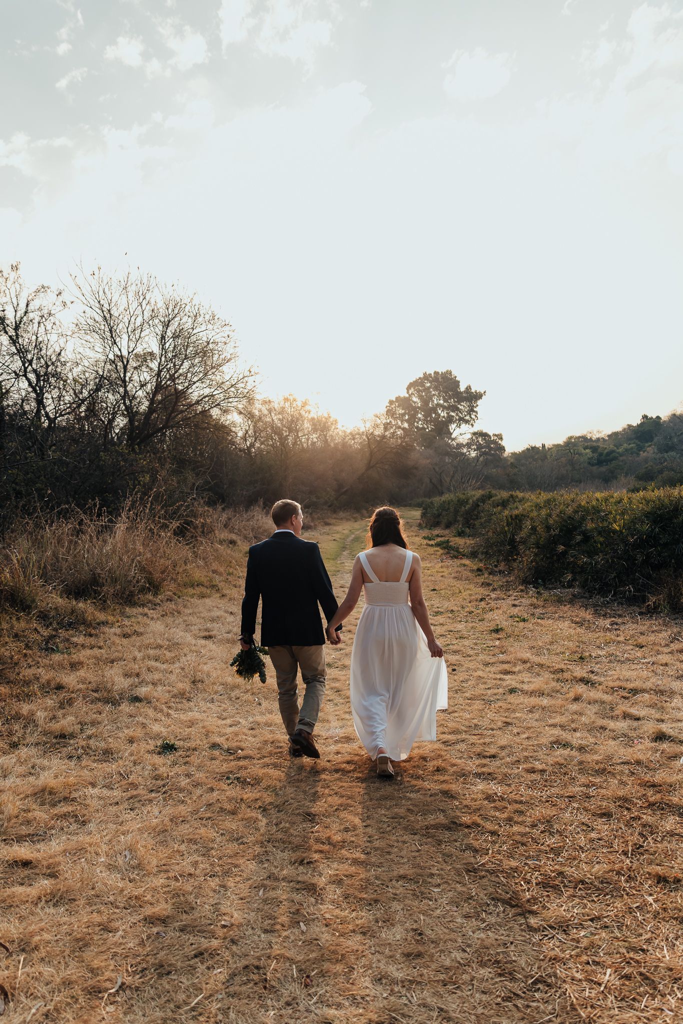 Couple walks through golden field during sunset with trees in background.