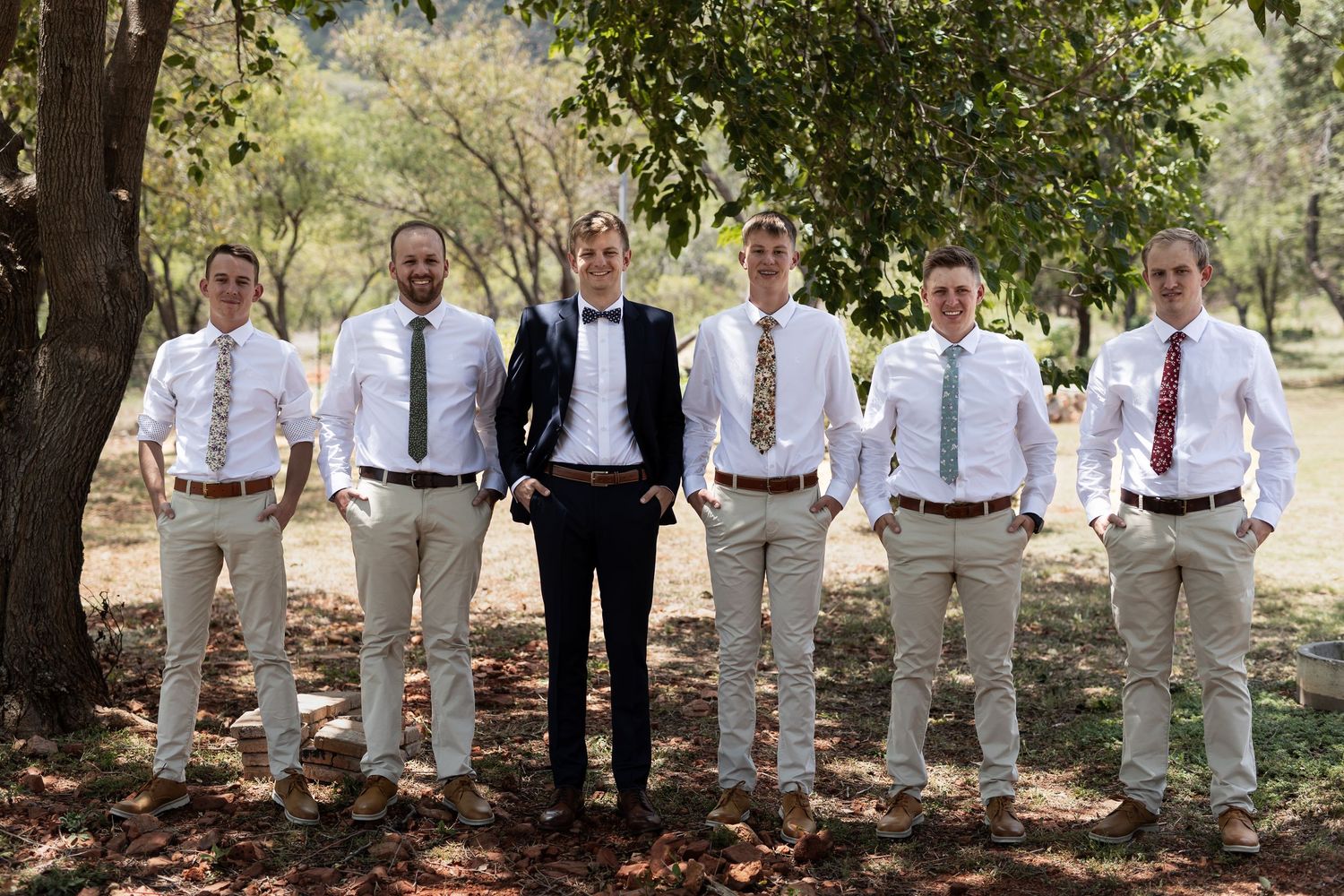 Wedding party in coordinated khaki and navy attire posed outdoors under trees.