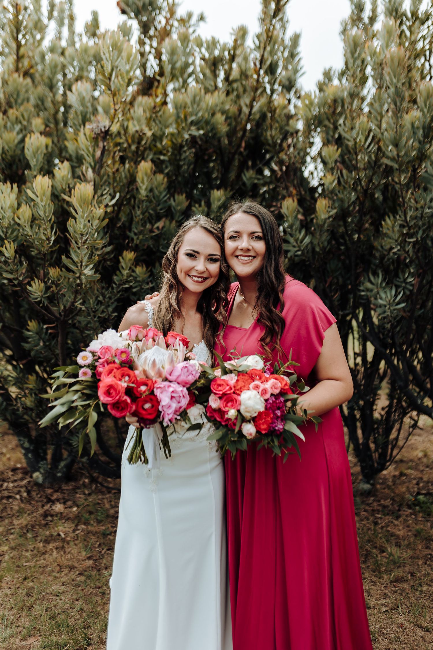 Bride in white gown and bridesmaid in red dress pose with pink and red bouquets outdoors.
