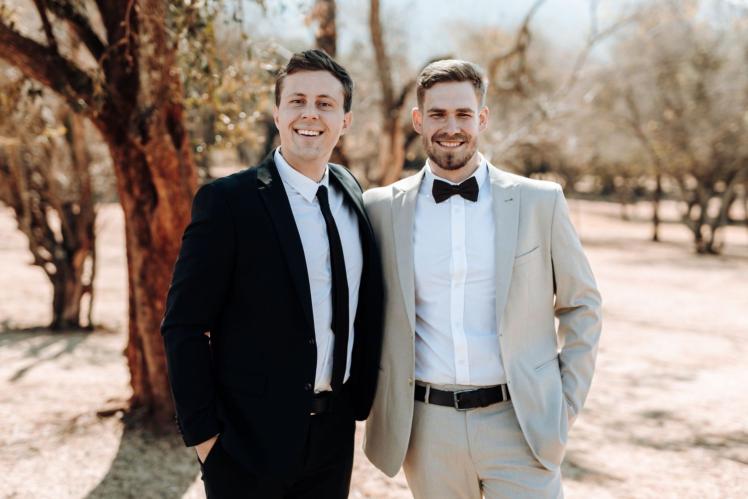 Two men in formal wedding attire pose for a photo in an outdoor desert setting.