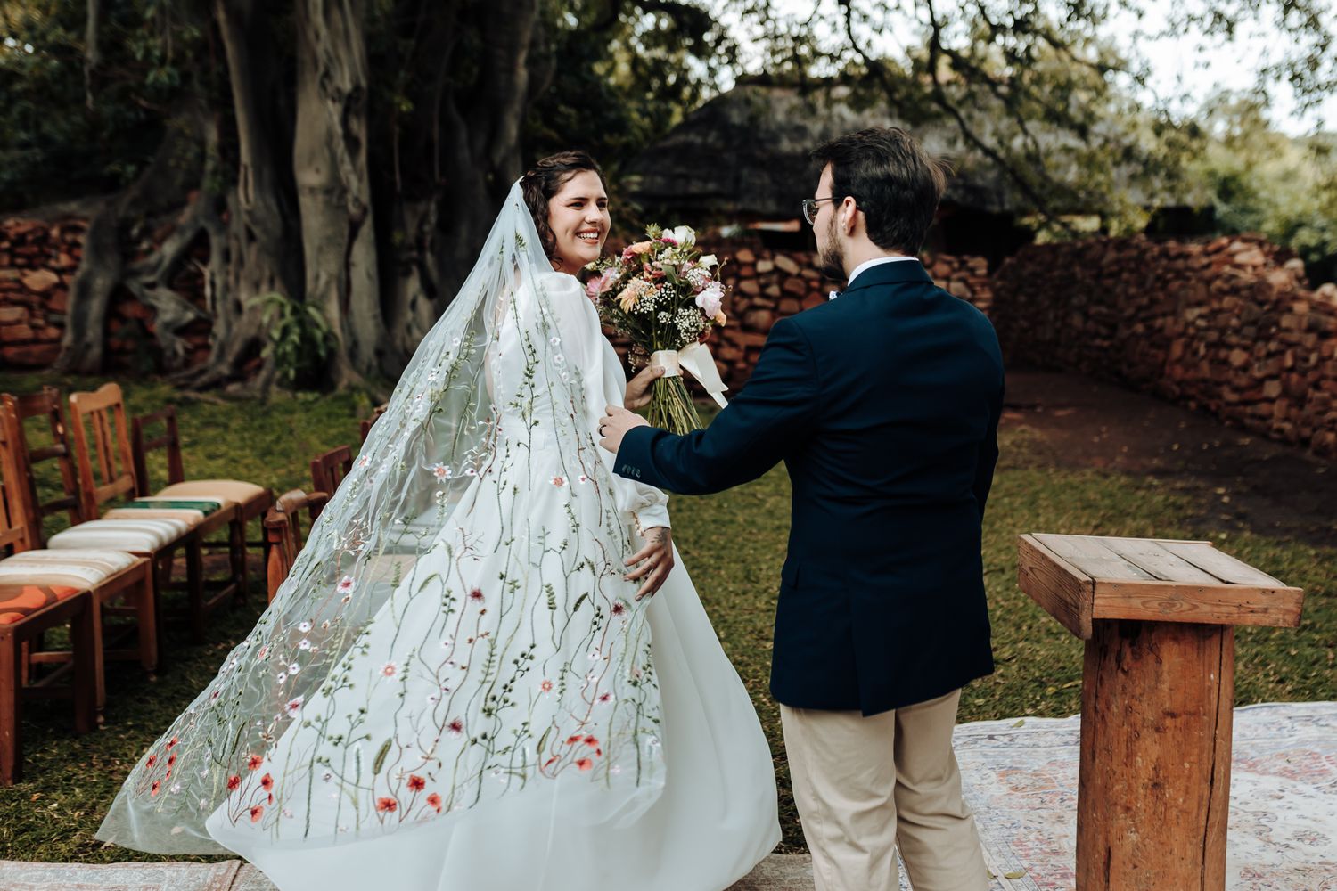 A bride in an embroidered wedding dress with a flowing veil at an outdoor garden venue.
