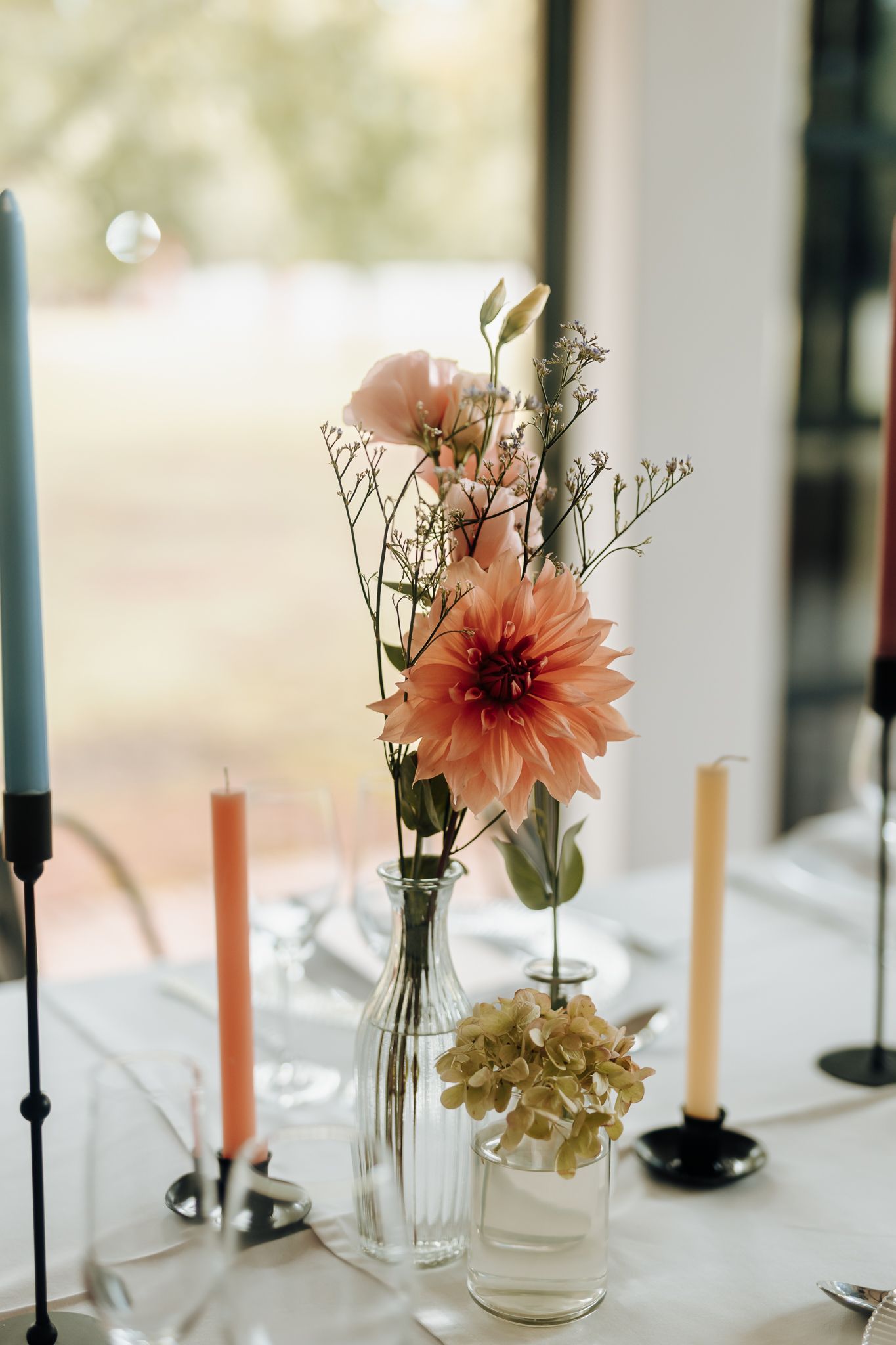 Simple floral arrangement with peach dahlia and delicate stems in clear glass vases with colored taper candles.