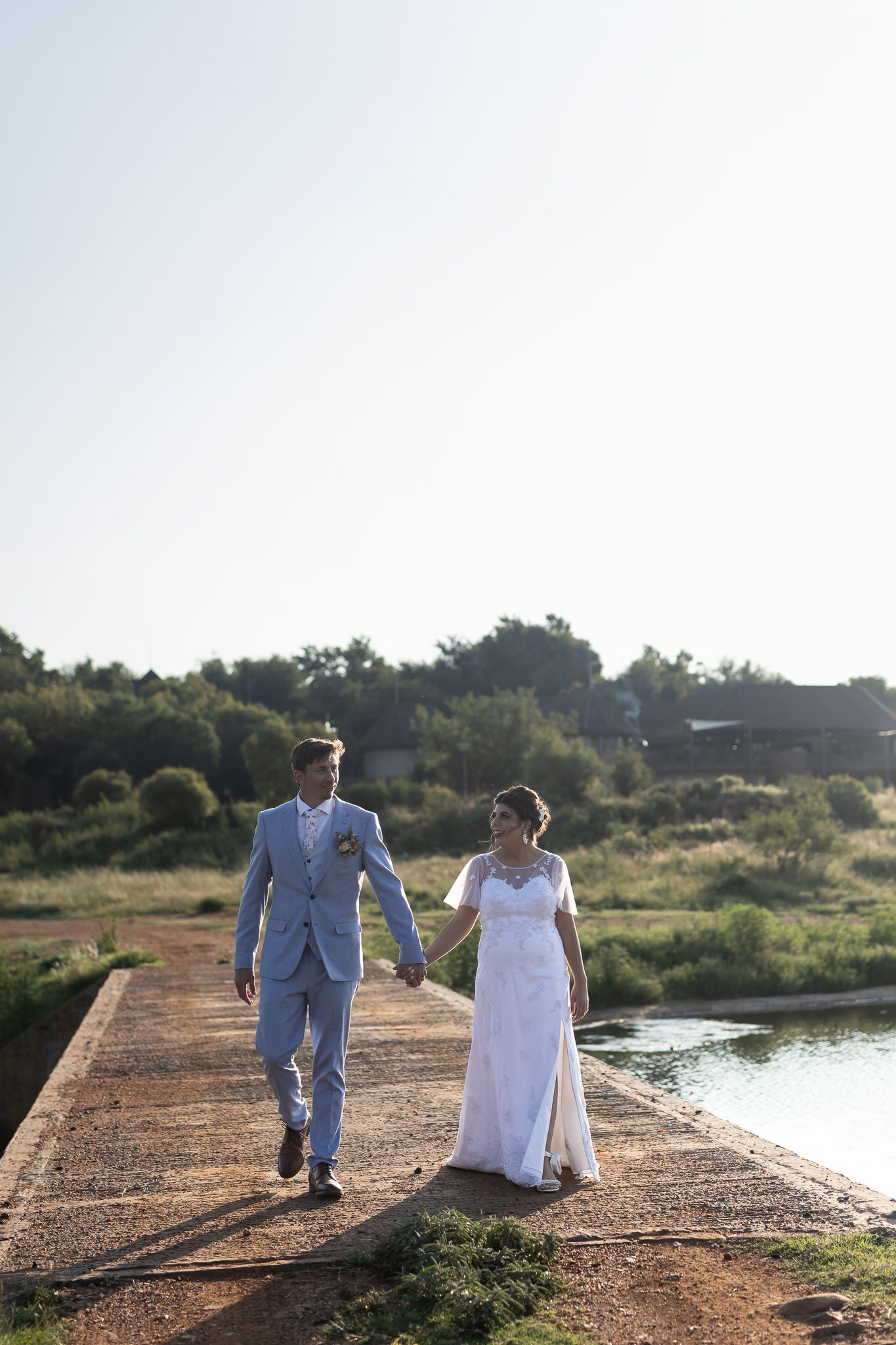 Newlyweds walking together on concrete pier with water and landscape in background.