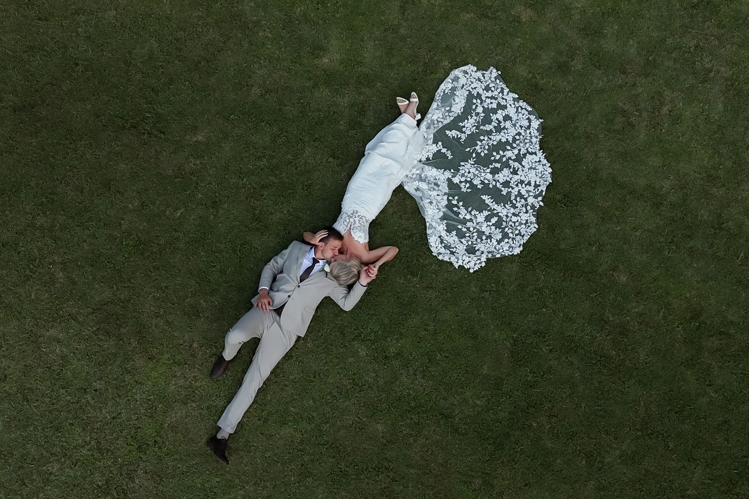 Overhead drone view of newlyweds laying on lush green grass with flowing white dress details.