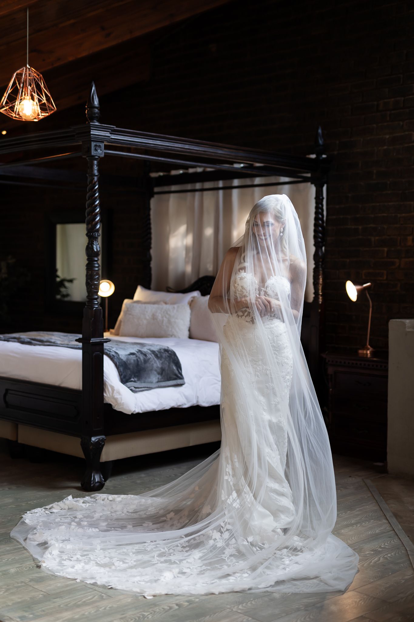Elegant lace wedding gown with long train displayed in a rustic bedroom with four-poster bed and exposed brick walls.