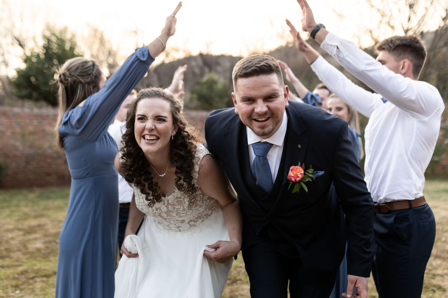 A joyous wedding party poses outdoors with energetic hand gestures and laughter in blue attire and traditional formalwear.
