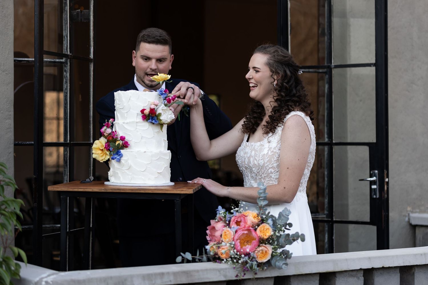 A couple cutting their floral-decorated white wedding cake together on an outdoor patio.