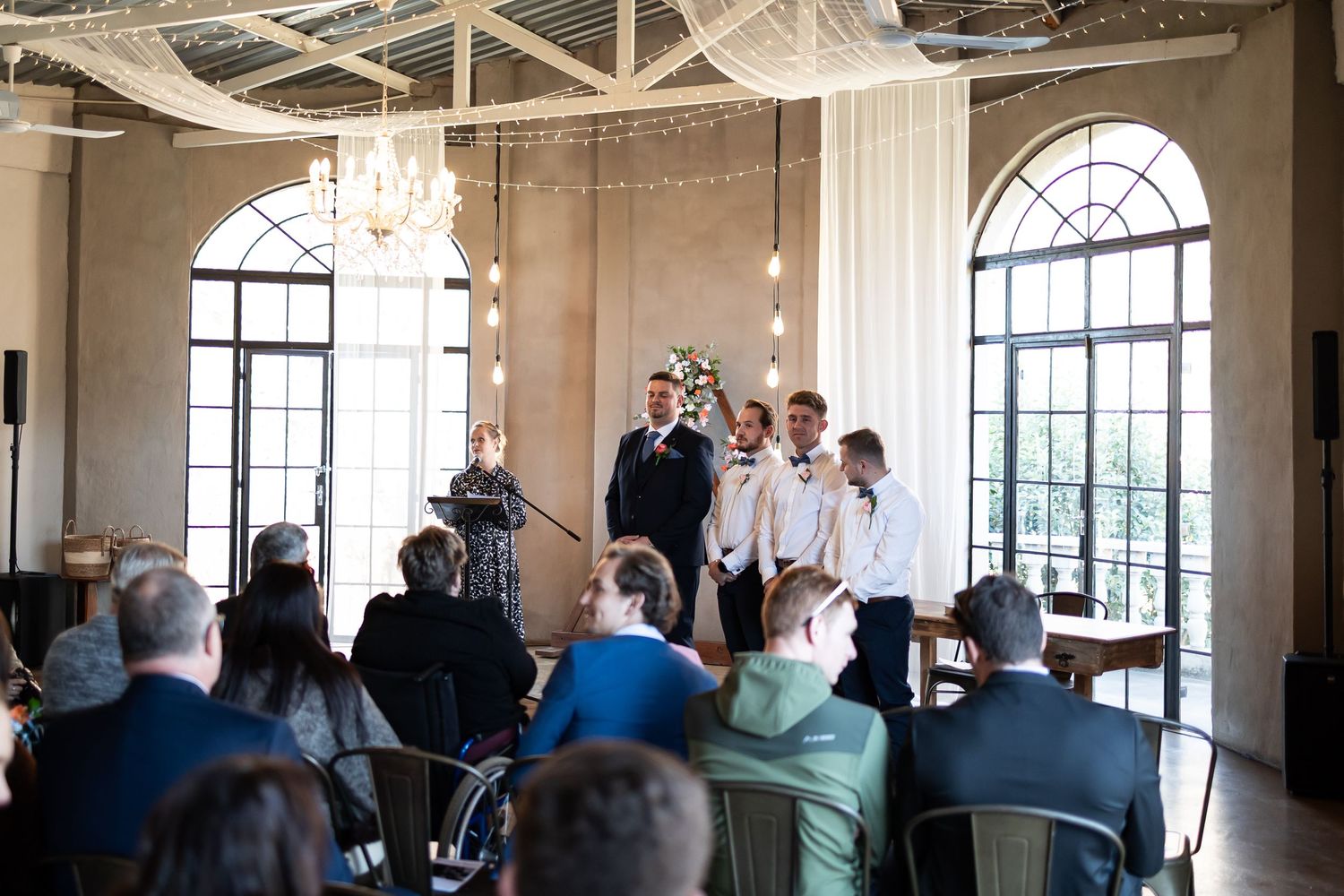 An elegant wedding ceremony takes place in a bright room with arched windows and a crystal chandelier.