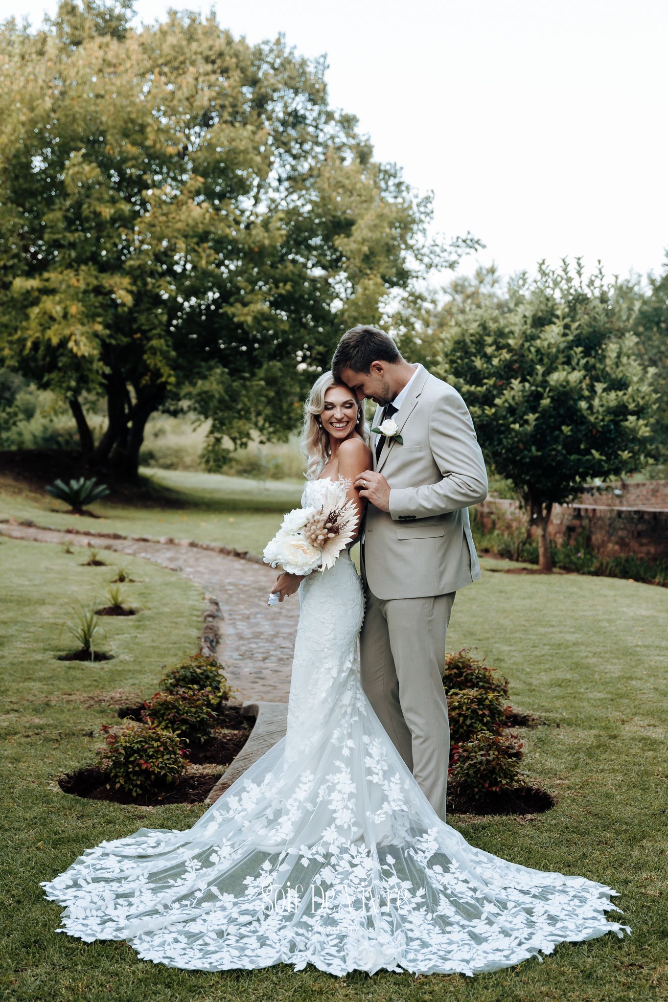Wedding couple embracing in garden setting with bride wearing stunning lace train dress.