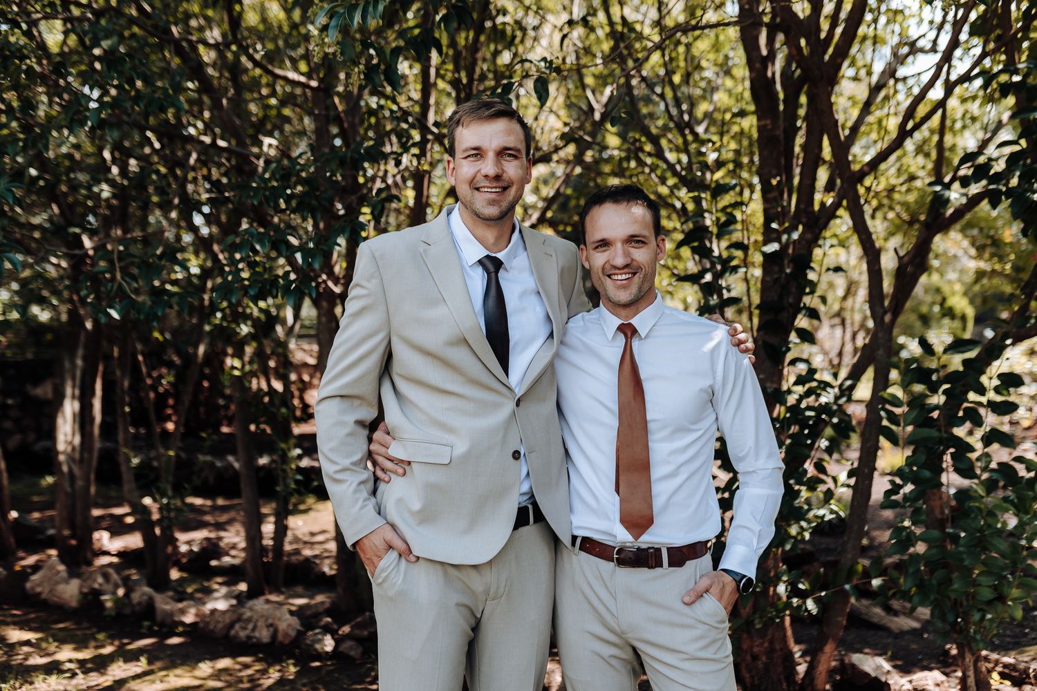 Two men in light suits and ties pose together for a wedding photo.