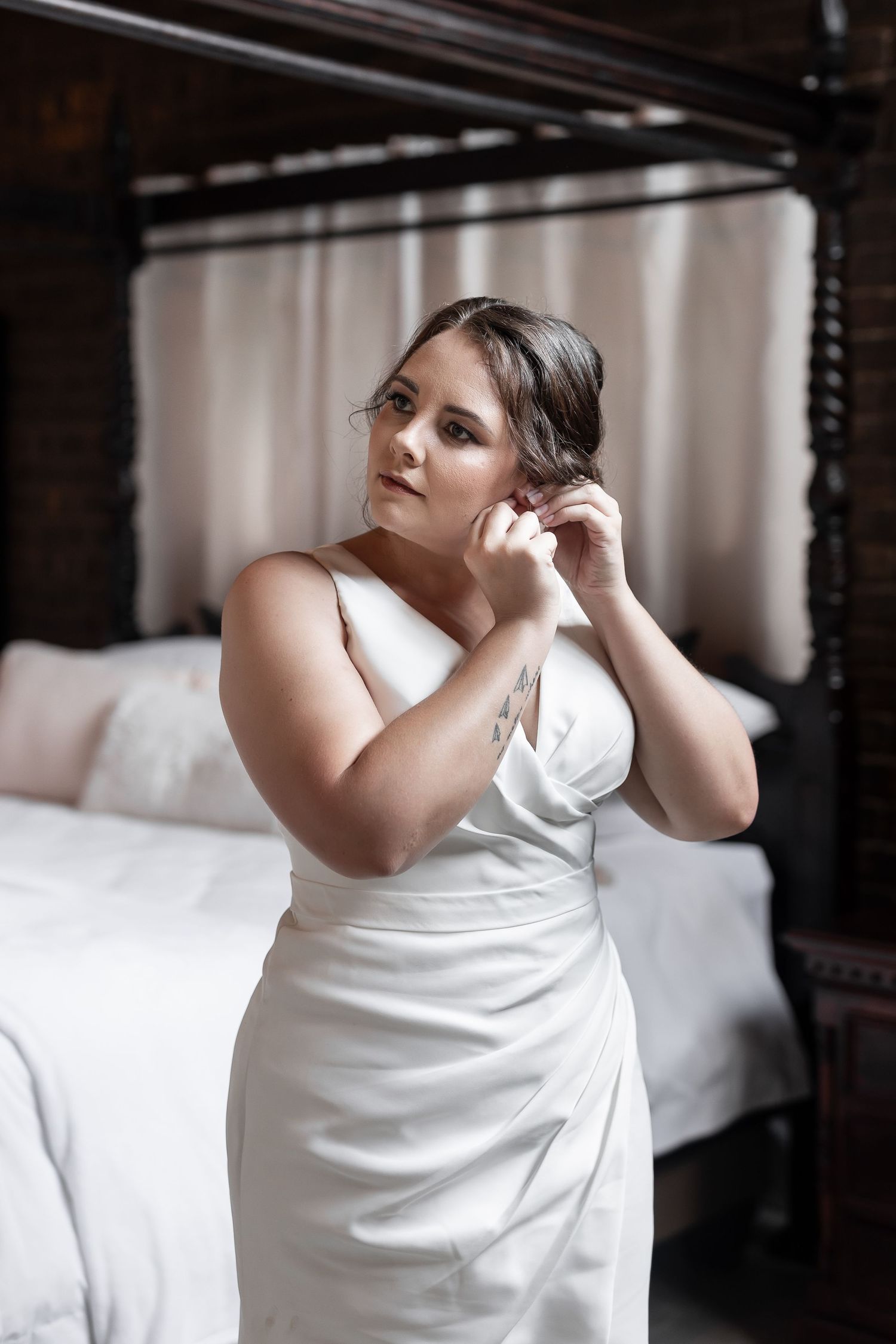 Bride in elegant white dress puts on earrings while getting ready in vintage bedroom.