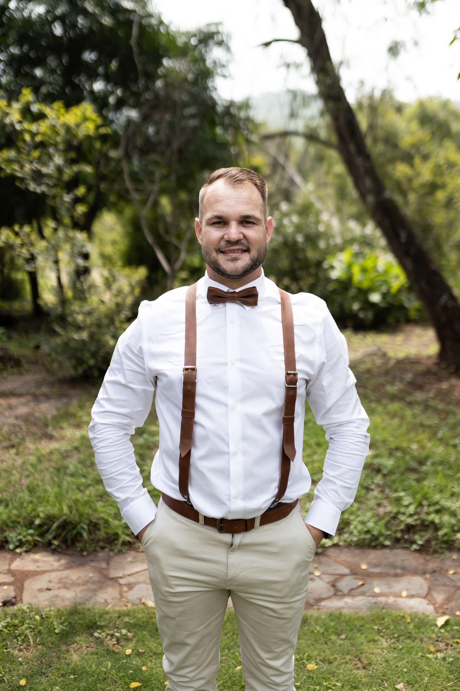 Formal portrait of groomsman wearing white shirt, bow tie and brown leather suspenders outdoors.