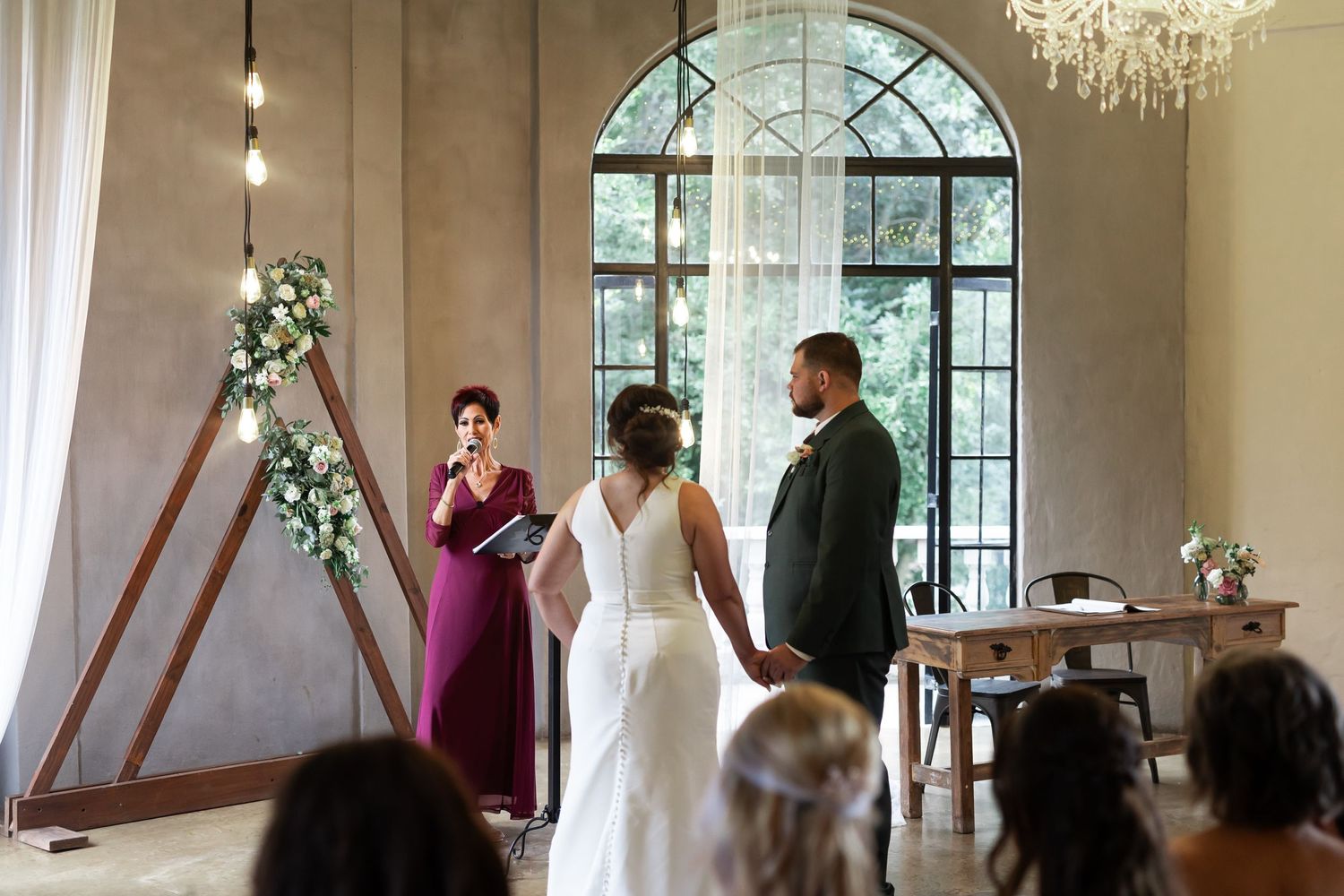 Wedding ceremony in bright venue with chandelier and arched windows.