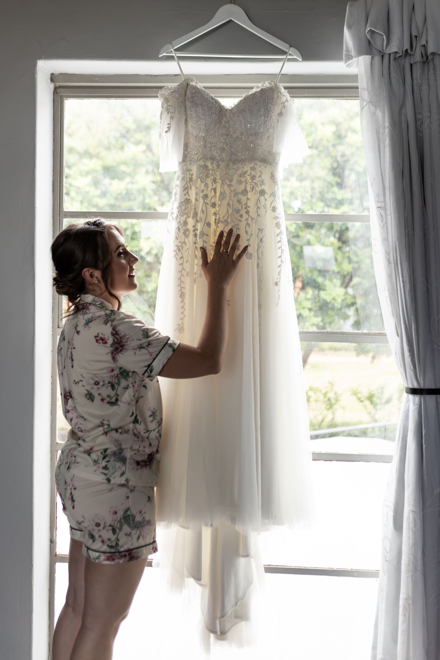 A white lace wedding dress hangs by a window while someone reaches to touch it.