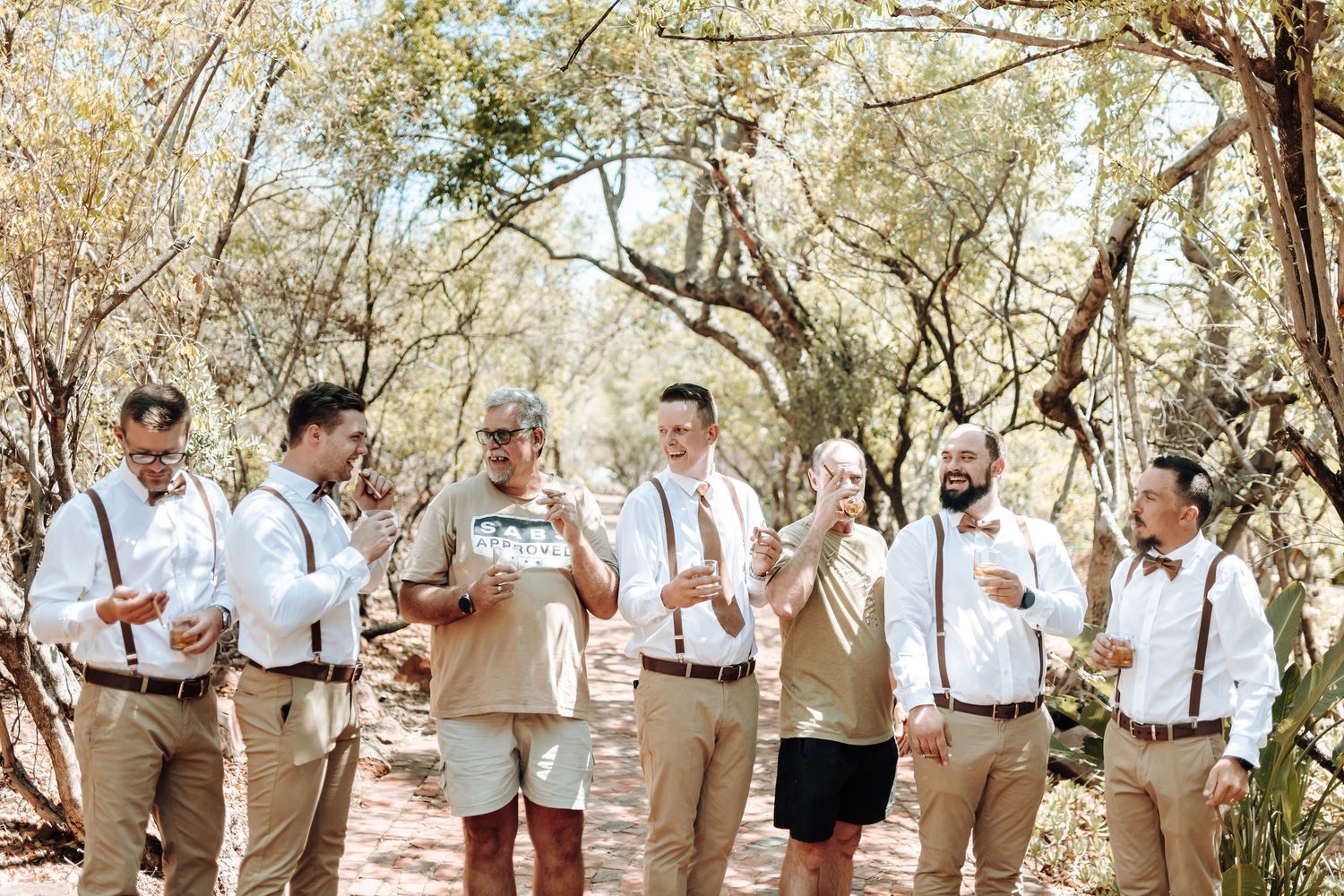 Groomsmen in suspenders and bowties enjoying drinks together under trees.