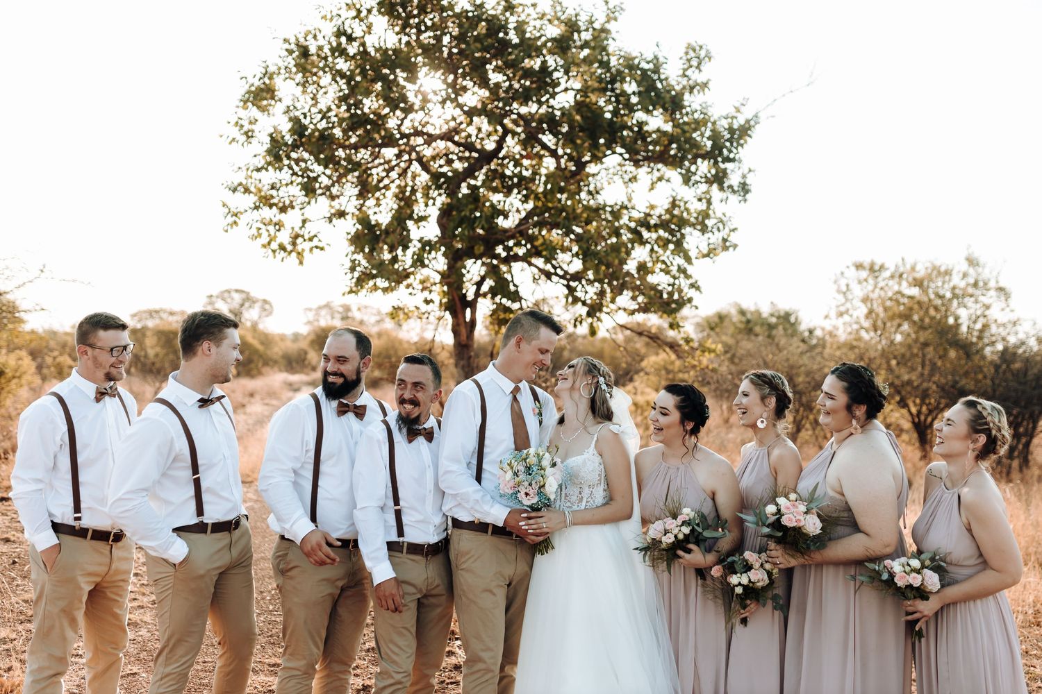 Wedding group photo featuring coordinated neutral-toned attire in sunlit outdoor setting.