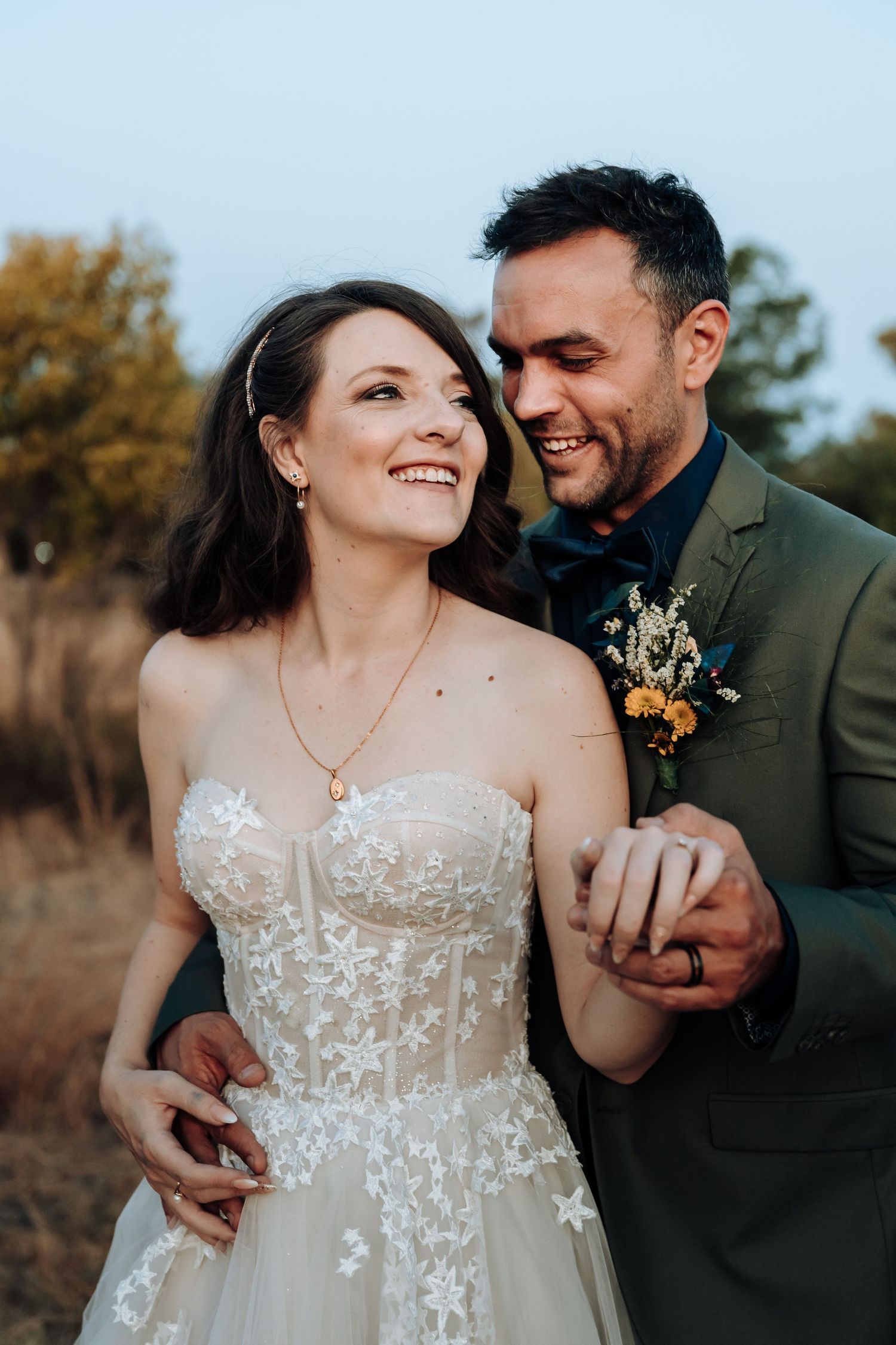 Happy couple sharing a joyful moment together outdoors with groom wearing green suit and bride in lace dress.