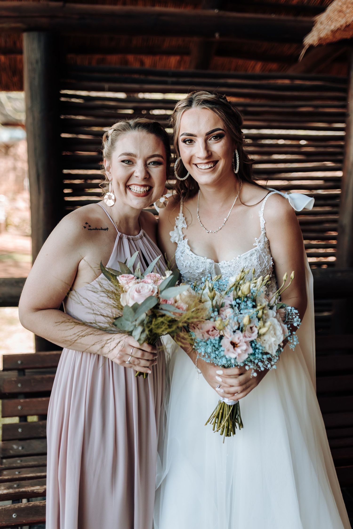 Joyful wedding portrait of bridesmaid and bride with matching bouquets under rustic roof.