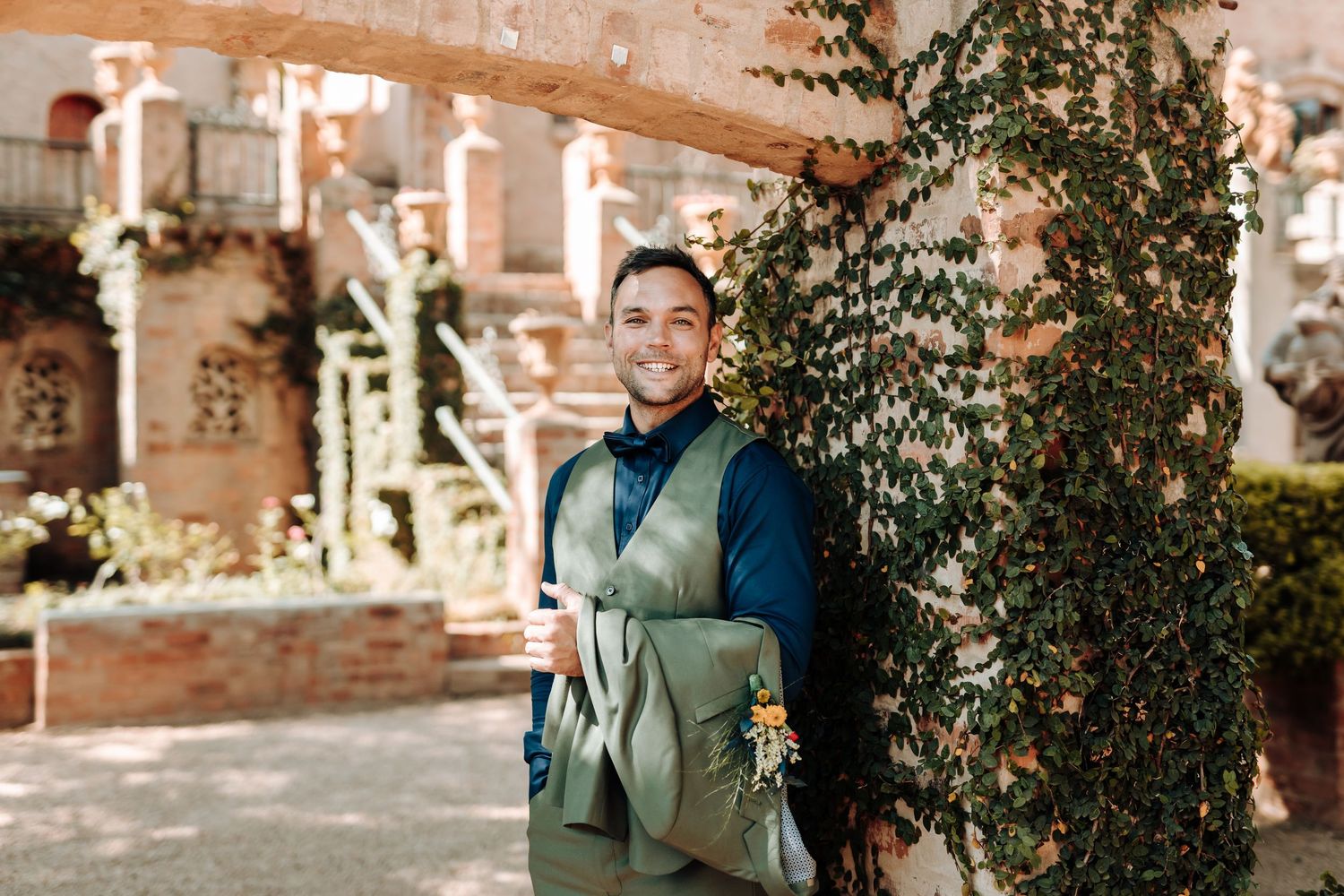 A person in formal attire poses outdoors near ivy-covered brick walls.