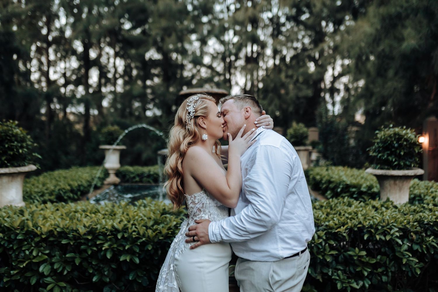 Newlyweds share a loving kiss in a formal garden setting with sculptured hedges.