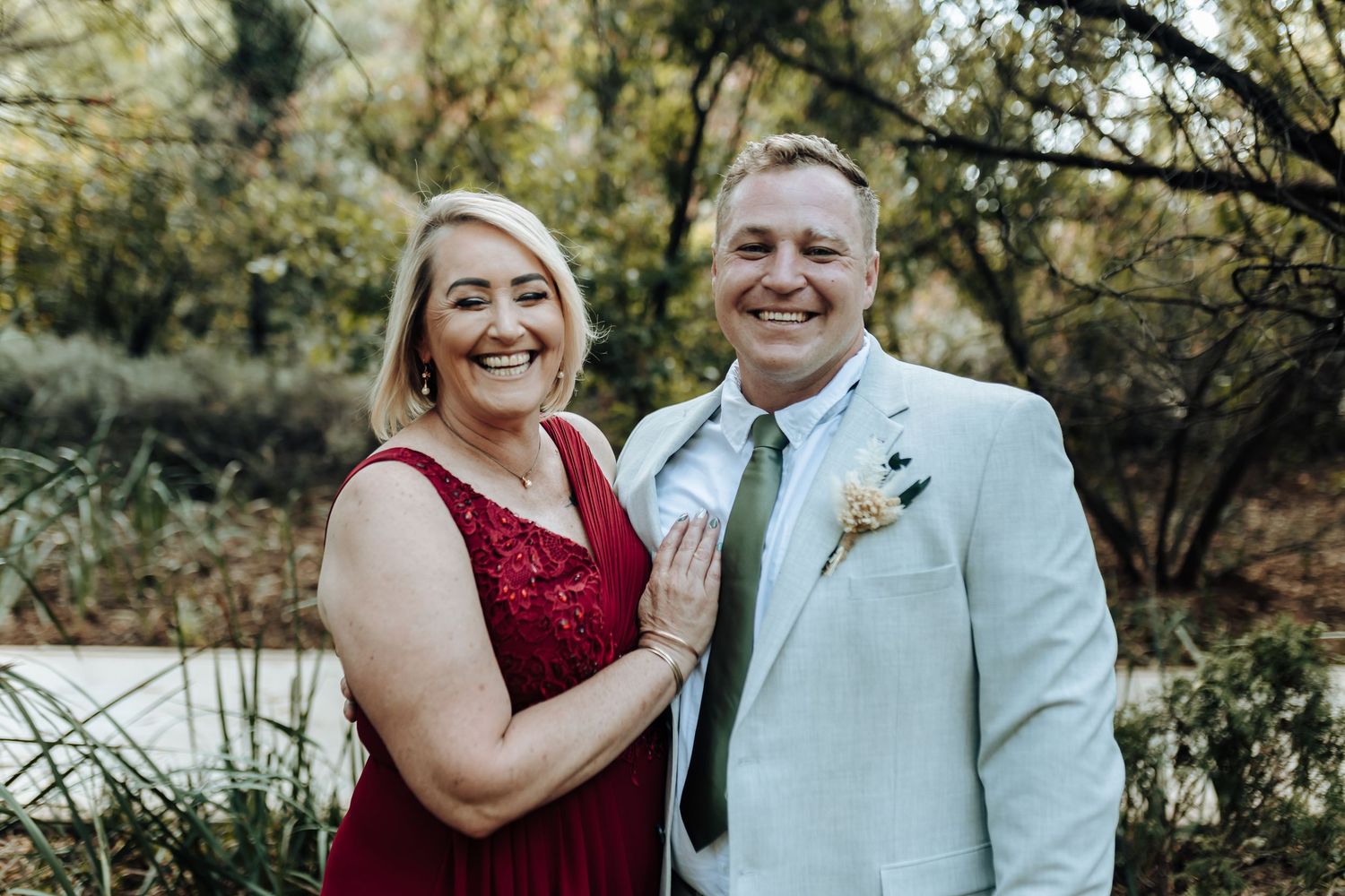 Couple in formal wedding attire smiling together outdoors with natural scenery.