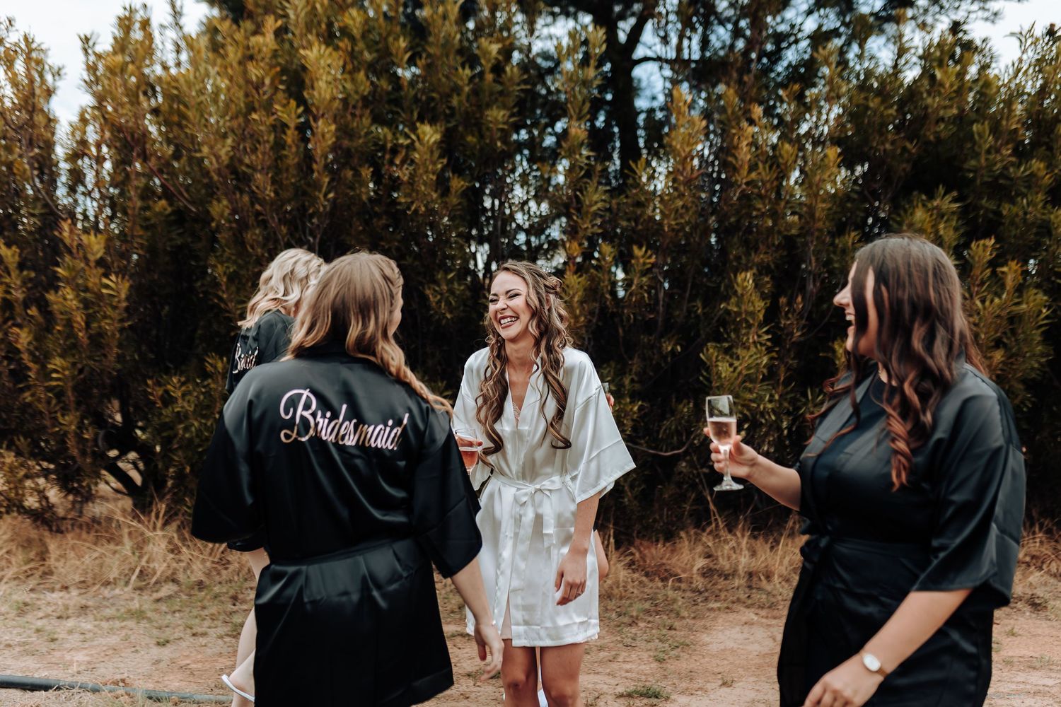 Bridal party in matching robes celebrates during a pre-wedding gathering outdoors.