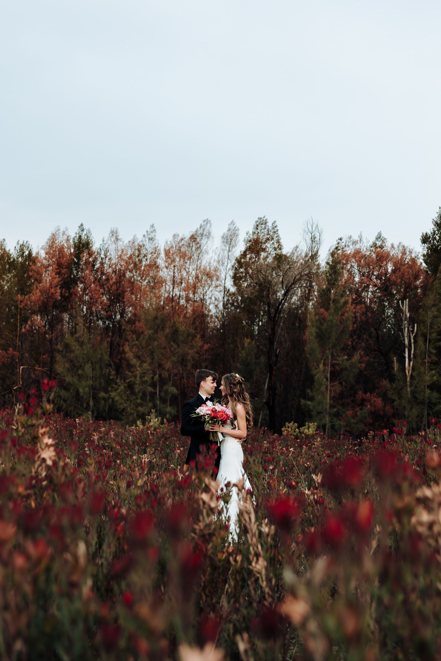 Wedding couple stands in dreamy autumn field with red wildflowers and forest backdrop.
