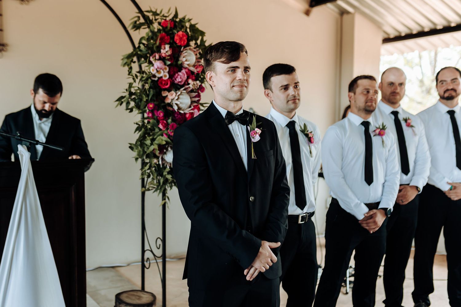Groomsmen line up at altar under floral arch during outdoor wedding ceremony.