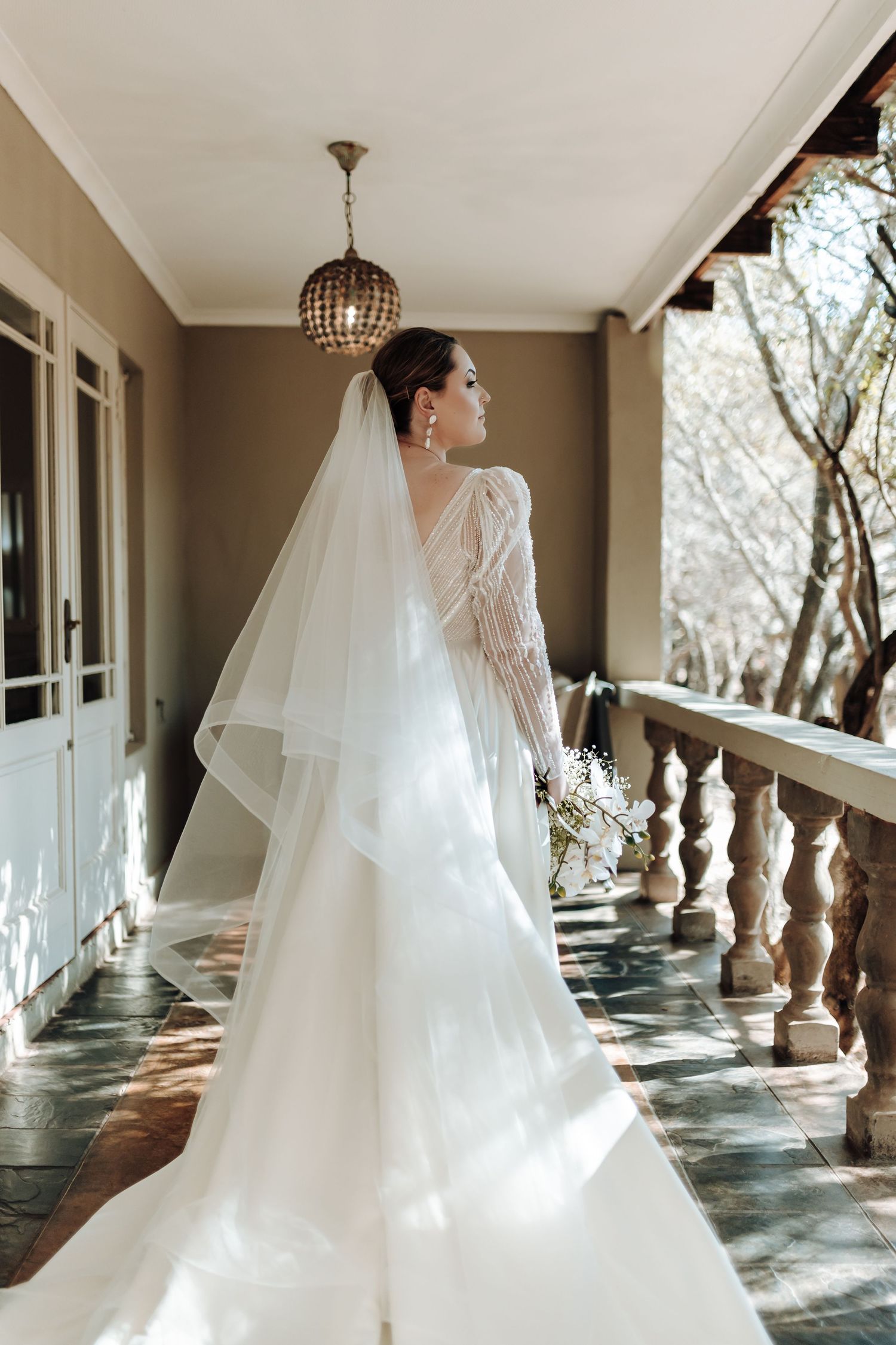 Bride in long-sleeved wedding gown poses on sun-lit veranda with cathedral veil.
