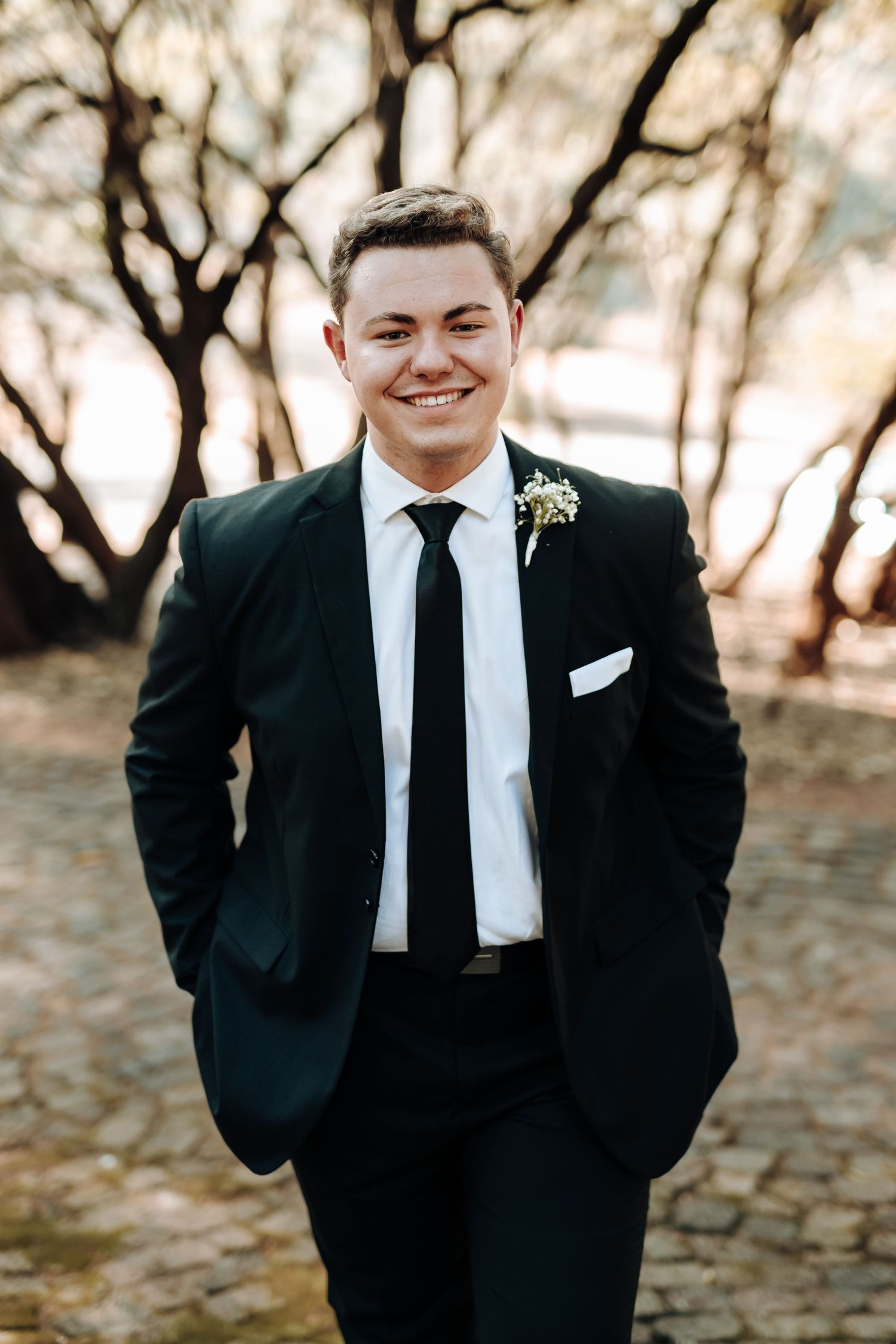 Groomsman in fitted black suit smiles brightly while posing outdoors.