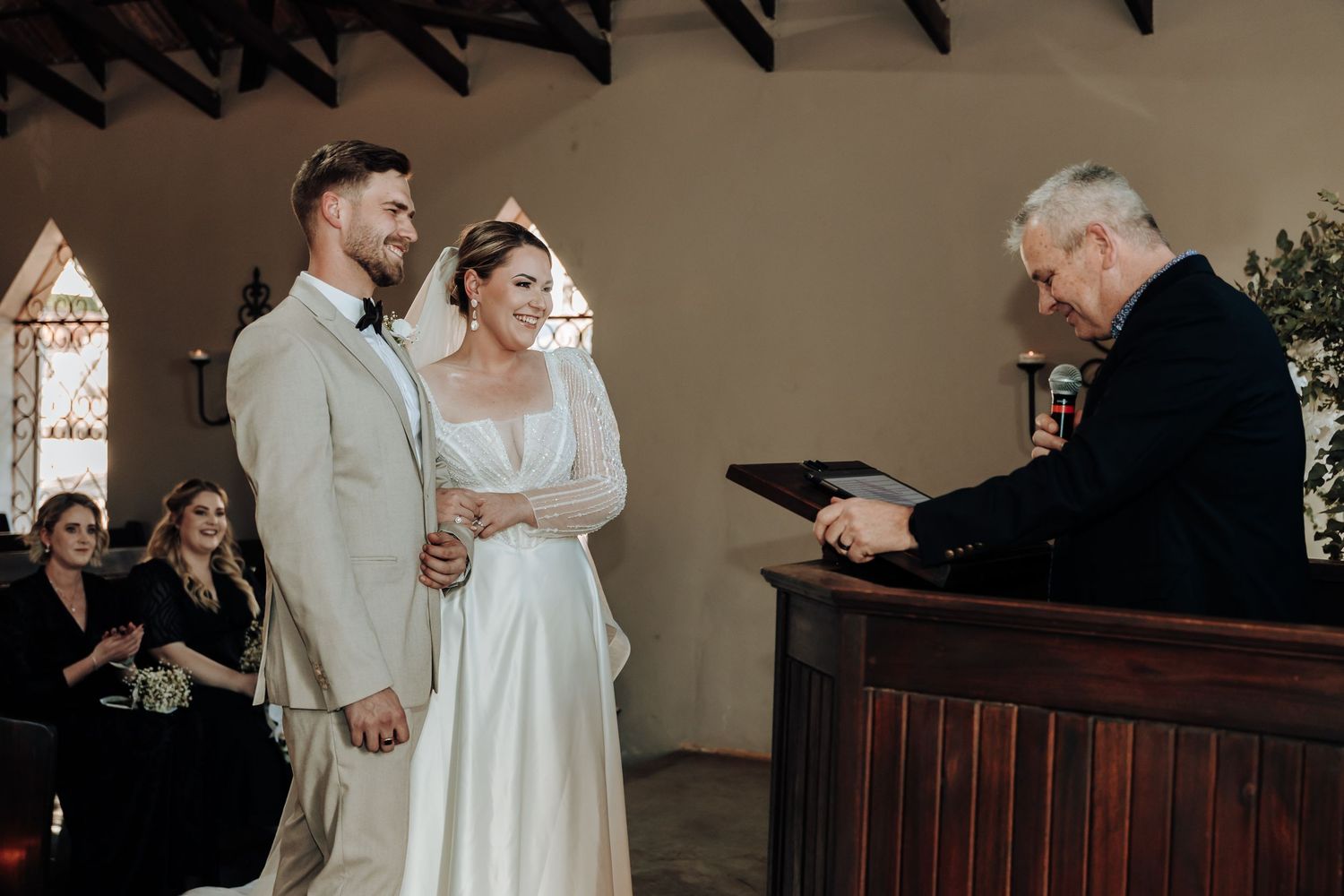 A wedding ceremony takes place in a traditional chapel as the officiant reads from a book.