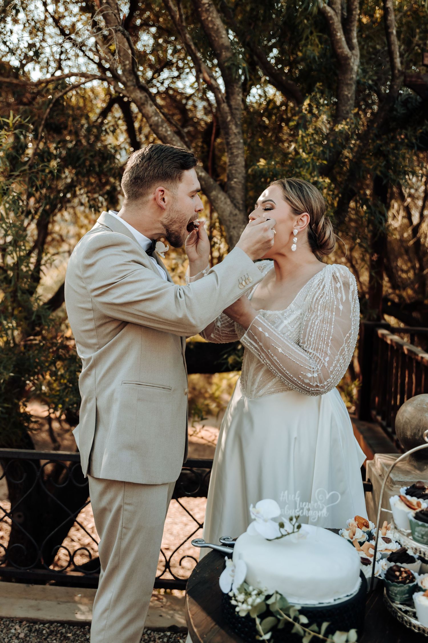 A couple shares a playful cake-feeding moment during their wedding reception in an outdoor garden setting.
